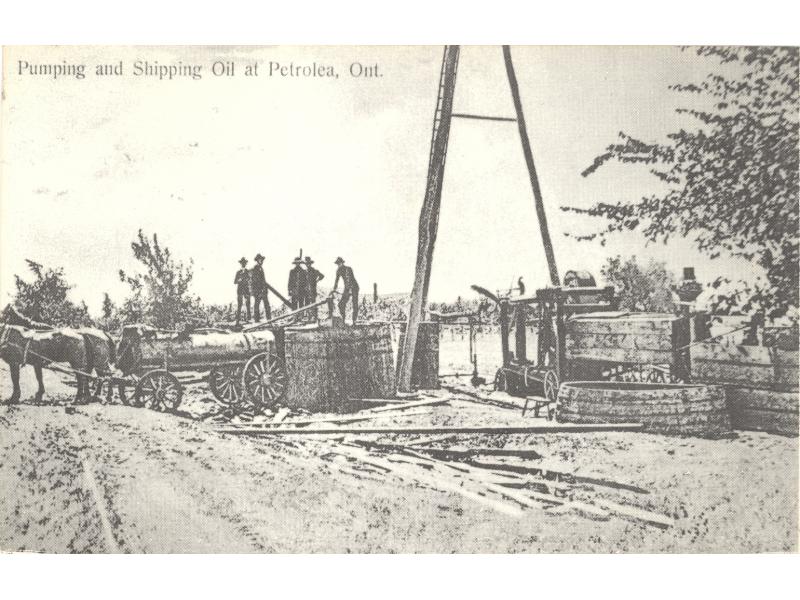 A group of men stands on top of a wooden storage tank beside a tank wagon pulled by two horses. There is a three-pole derrick behind them.