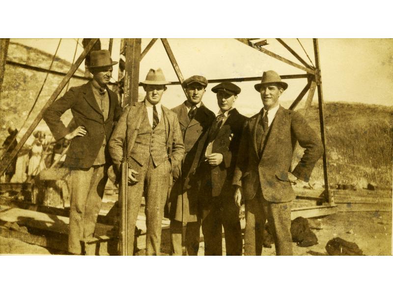 A photo of five International Drillers standing at the base of an oil derrick. They are wearing suits and hats.