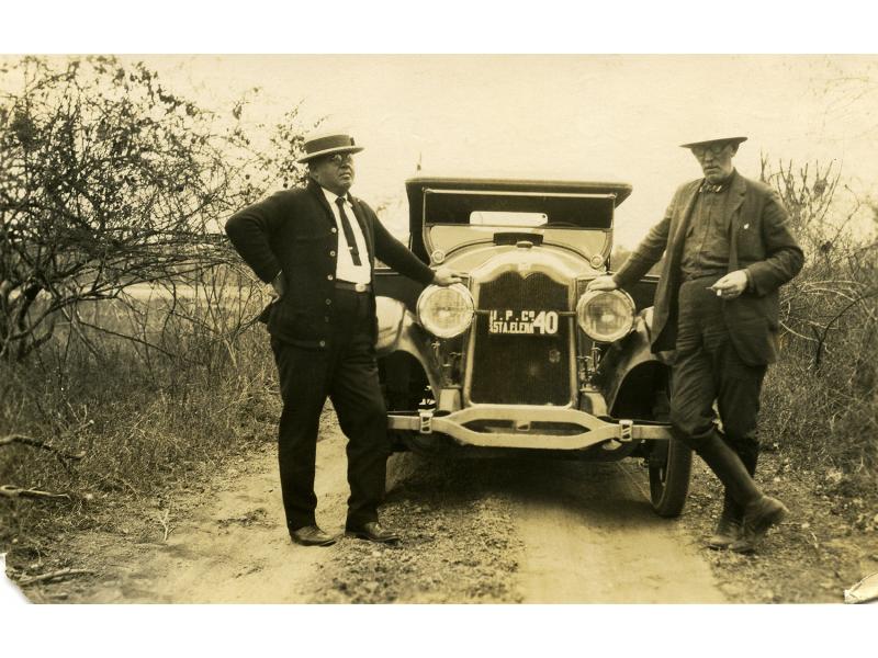 Webb and Burns standing on either side of a car parked on a dirt road. There are short trees on either side of them. The licence plate between the headlights reads: 1924. I.P.Co. STA.ELENA. 40".