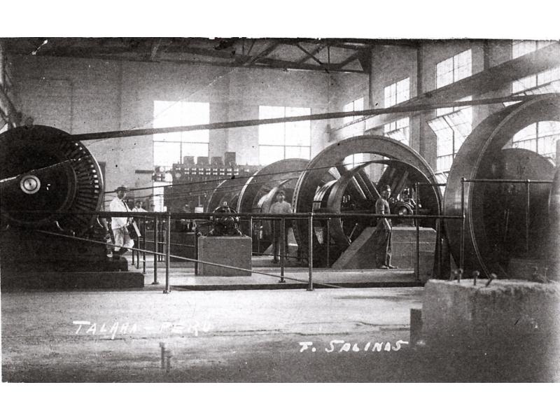 Inside a refinery with four large wheels attached to smaller wheels with belts of fabric. Men stand near the wheels. There are windows letting light into the room.