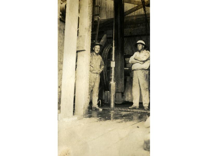 Two men wearing work clothes and hats standing inside a drilling rig. There is a large pipe and a wheel in behind them.
