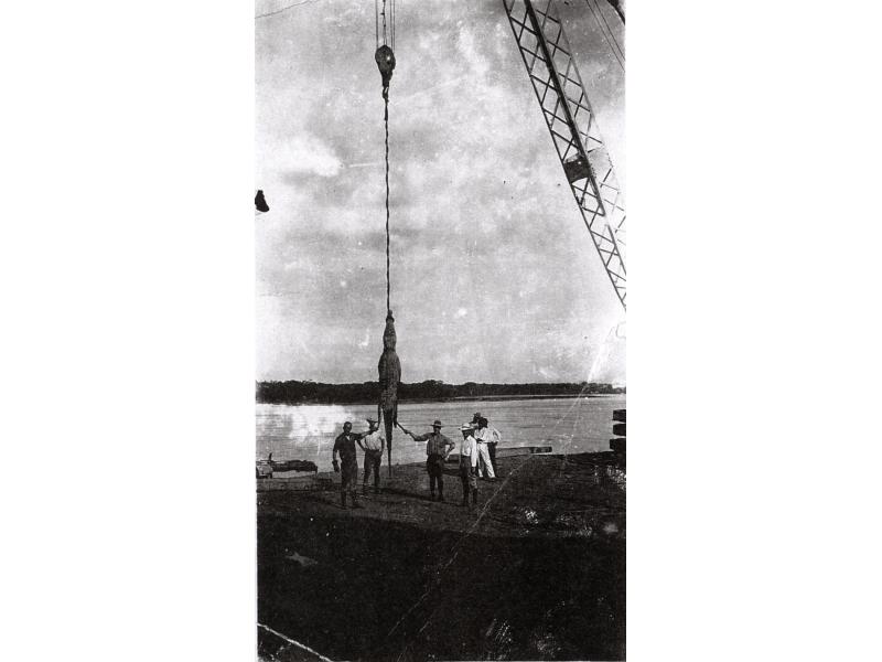 An alligator suspended from a crane, with four men standing around it. There are two people in the background. They are beside the edge of a lake or river and there are trees on the other side of the water.