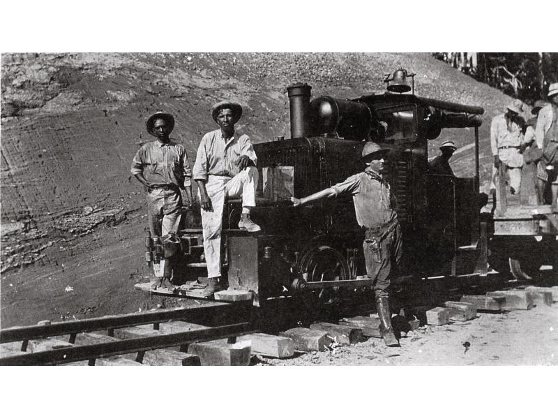 Three men stand at the front of a train engine with a bell on top of it. There are men standing on a cart attached behind. The railway tracks are raised off of the ground with wooden blocks. There is a dirt hill in the background.