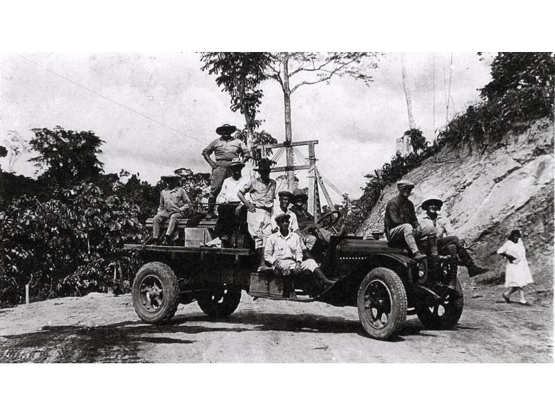 Eight men sit on a long car parked beside a hill. A woman walks behind them. There are trees in the background.