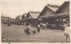 Front of a postcard showing three identical wooden buildings with two peaks in the roof. There is a line of carts in front of them. There are people walking around in an open space and a man is carrying two baskets connected by a pole on his shoulder. 