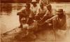 A photo of a group of men crouching with an alligator on a river bank. Its mouth is open, showing its teeth. 