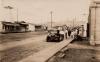 A photo of a dirt road with a wooden sidewalk running beside it. There is a group of people near a car parked on the side. There are single-story buildings on either side and hills in the background. 