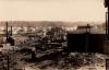 A photo of an oil refinery. There is a dark storage tank with stairs leading to the top on the right. There are buildings with pipes running between them in the middle and a group of light-coloured storage tanks at the back. There are hills in the background. 