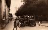 A photo of two cars parked side by side on a cobbled street. There is a boy on either side of the two cars and a building to the left with a woman walking past the corner. A group of trees is to the right and there is a white tower down the road. 