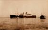 A photo of a ship with two masts and two smoke stacks, anchored in the water. There is a smaller boat just off to the left. There are people getting into small boats at the base of the large boat. 