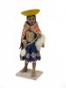 Front view of a figurine of a woman wearing sandals and who is dressed in a blue skirt with an embroidered design along the edge, as well as an orange rectangular shoulder cloth fastened at the front. She has an inverted yellow hat and is carrying a spool and wool. 