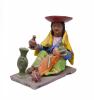 A figurine depicting a woman wearing a green skirt, a purple blouse, a blue vest with a design on it, and a yellow rectangular shoulder cloth. She is wearing an inverted red hat. There is a baby in her lap and she is holding pieces of pottery. 