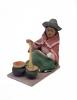 Figurine of a woman stirring two black pots with a brown spoon. She is wearing sandals, a green skirt, and a red rectangular shoulder cloth. There is a white apron wrapped around her wrist and she is wearing a grey hat with the brim curled up. 