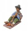 Figurine of a man playing the flute wearing sandals, green pants, a blue shirt, and a brown poncho over one shoulder. He has a grey hat and there are three orange egg-shaped objects at his feet. 