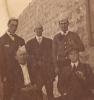 Five International Drillers, three standing and two sitting. They are wearing dark suits. Four of the men are wearing paper hats and one (bottom right) is wearing an ascot cap.  