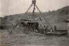 A group of men are putting together a bulldozer using three poles tied together at the top with a chain coming down from the middle to help lift the heavy pieces. There is a hill dotted with trees in the background. 