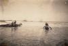 Three men standing on a raft made from logs and three men in a canoe. They are in shallow water and there are small waves moving towards them. There are two ships out on the water in the background.