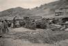 A photo of a half-assembled bulldozer. George Rawlings stands to the left. There are stacks of pipes in behind the bulldozer and two buildings on the hill at the back of the scene.