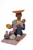 A figurine showing a man sitting on a pink mat and feeding a baby. He has an inverted orange hat and is wearing sandals, blue pants, blue shirt, yellow vest, and green poncho thrown over one shoulder. There is a vase by his right foot.