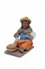 Figurine of a woman spinning wool wearing sandals, an orange skirt, a white apron, a blue blouse, and a brown rectangular shoulder cloth. She has a yellow hat and there are two brown bowls by her feet.  