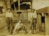 Three men in light shirts and dark pants, holding guns, in front of a wooden picket fence. The man in the centre is sitting on the ground and the other two are standing. 