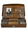 An open leather case with a metal latch on the front and removable organizers. There is a photo of a man in a suit along with scissors and other tools in the top part of the case. There is a belt across the bottom section. 