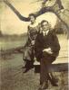 A woman in a long, dark dress and a man in a dark suit and tie sit on wooden planks beneath a tree branch.