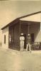 A photo of two International Drillers standing beside a white house with dark wood along the edges and railing of the porch. They are wearing white pith helmets. One has a white shirt on and the other a dark jacket. 