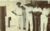 A photo of four men dressed in white on the deck of a ship. They are standing in front of a metal column with rivets in it.  