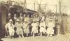 A photo of two rows of people on a wooden deck. The men are standing in the back row, wearing suits. The women are sitting in chairs in the front, wearing dresses and hats. 