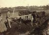 A photo of a man in a suit holding a wooden plow attached to two skinny cows. They are standing in a field. There is a man in a long robe standing behind him. 