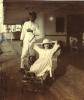 A woman lying back in a wicker reclining chair, wearing a white dress and hat. A man in a white suit stands behind her. They are on a ship deck with a wooden floor.