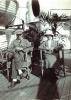 A man and a woman sitting on the deck of a ship. There are life boats hanging to the left and a palm frond behind them. The floor is made from wooden boards.