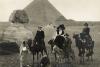 Une carte postale montrant quatre personnes chevauchant trois chameaux et un âne devant le Sphinx et une pyramide. 