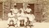 A photo of three men standing and four men sitting in front of a stone building with two screen doors and a window. They are wearing white and various types of hats.