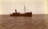 A photo of a ship on camp water. It is listing slightly to the side and the anchor is down to the right. There is one mast and a smokestack.