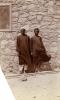 Une photo de deux hommes portant des robes foncées, debout devant un mur de briques avec une fenêtre munie d’une moustiquaire.  