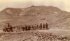 A photo of Egyptian workers standing in a line, hauling a boiler with ropes along a road in the desert. The boiler has four weeks and is has two cylindrical components put together. 