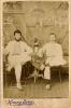 A photo of two men wearing white, sitting cross-legged on chairs on either side of a small table covered with a cloth. There is a vase of flowers on the table. Both men have a cane in their right hand and are balancing pith helmets on their left knee. There is a painted background of temples in the background.