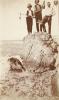 A photo of three men stand on top of a large rock. The man on the right is holding a fishing spear, the man in the middle a gun, and on the left a survey rod. The tide has gone out and mud can be seen under a shallow layer of water.