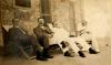 A photo of four men sitting in reclining chairs in front of a stone building. The two on the left are wearing dark jackets and the two on the right are in white.