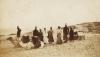 A photo of seven Egyptian men standing with a white woman a desert. There are two camels with saddles on their backs sitting in front of them.