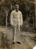 A photo of William O. Gillespie in a white suit with a dark bow tie and shoes. He is standing on a path in front of trees. 