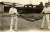 Irwin and Lovelady holding a boa constrictor in between them. They are standing on a dirt road and there are buildings behind them. They are wearing white shifts and pants. 