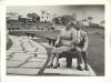 A photo of Vola and John Braybrook sitting on a park bench. There is a path with large stones beside them and a series of benches along the path behind them. She is wearing a skirt and jacket with high heels and he is wearing a suit and tie.