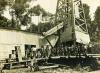 A group of men standing in front of an oil rig that is covered in sheet metal. There is a pile of casing in front of them and there are trees in the background. 