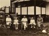 Three men and two girls (alternating) sit in chairs outside a building on stilts. They are wearing light clothing. The building has a porch with stairs and white and dark railings and posts.  