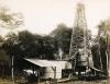 A photo of five men standing by a pile of casing in front of a drilling rig. There is a structure with a roof in the foreground, and a line of trees in the background.