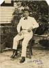 A photo of Albert Haley sitting on the arm of a chair. He is wearing white pants, a white shirt, a bow tie, and a hat. There are bushes behind him as well as a building with white siding.