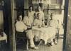 A photo of seven International Drillers around a table indoors with a white tablecloth. They are wearing white and four are sitting on wicker chairs. There is a cup, a pipe, and a bottle on the table.